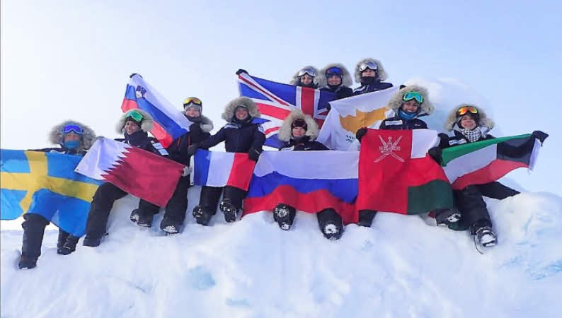 The skiing group is sitting on a mound of snow at the North Pole holding up their respective national flags